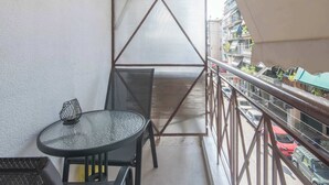 Een balkon met een glazen tafel en stoelen, een metalen scherm en uitzicht op geparkeerde auto’s en gebouwen.