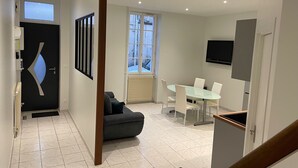 A modern living room with a sofa, a dining table with chairs, a TV mounted on the wall, and a large window with a view of the outside. - Logis du 96 (Angoulême)