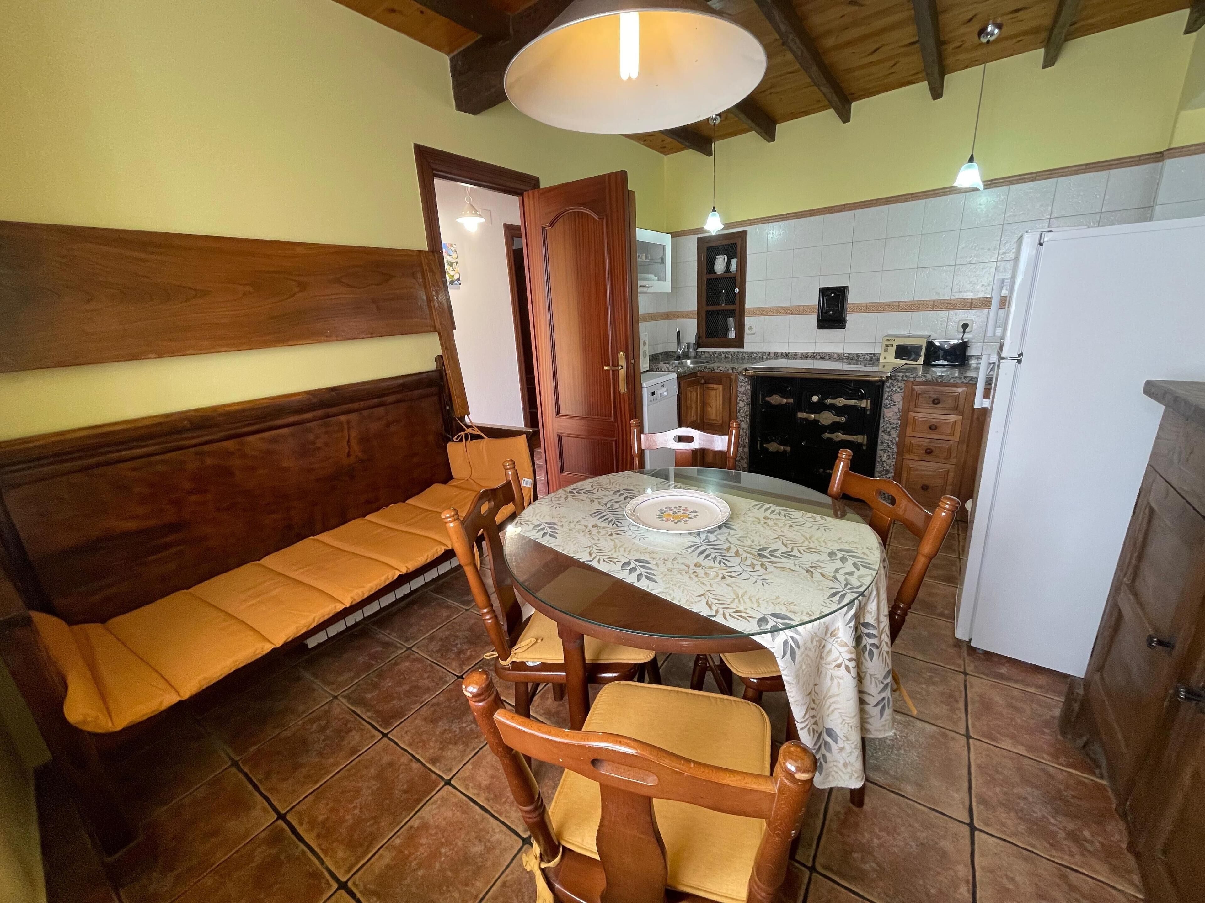Un espace repas avec une table ronde, des chaises, un banc, une cuisine en arrière-plan et un plafond en bois.