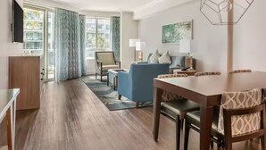 A modern hotel room with a wooden floor, a dining table with chairs, a sofa, a TV, and a window with curtains. - Ocean Oak by Hilton Grand Vacations: Your 2-Bedroom Coastal Haven (Hilton Head Island)