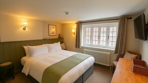 A hotel room with a bed, a wooden table, a chair, a radiator, a window with curtains, and a wall-mounted TV. - The Eight Bells Inn (Bolney)