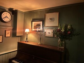 Una habitación con un piano, un reloj de pared, cuadros enmarcados, un candelabro con velas y un florero con flores.