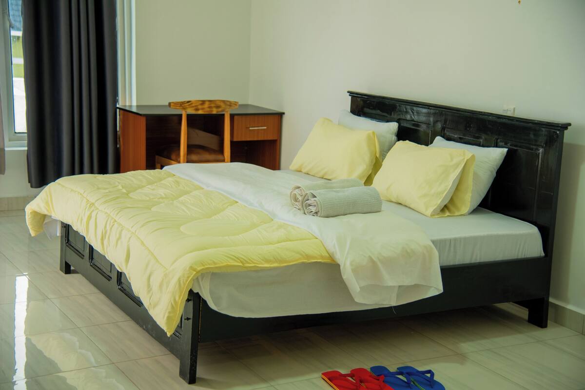 A neatly made bed with a yellow duvet and white linens, a wooden nightstand with a lamp, and a window with curtains.