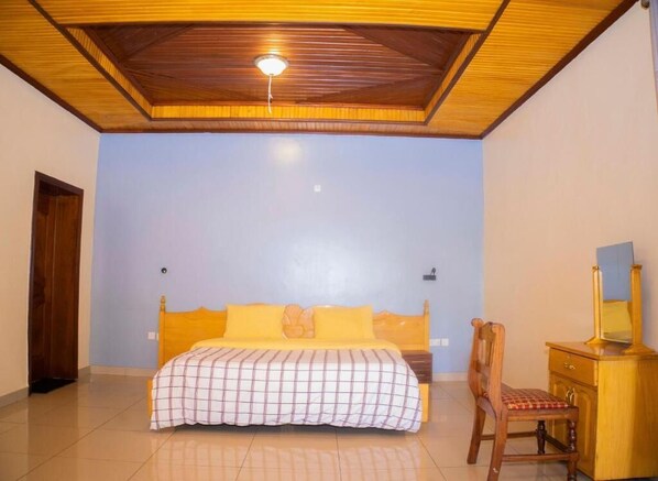 A bedroom with a wooden ceiling, a bed with a checkered bedspread, a wooden chair, and a wooden dresser with a mirror. - Kibagabaga Comfort Stay (Kigali)