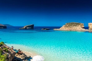 A coastal beach with clear turquoise waters, rocky formations, and a few people enjoying the view.