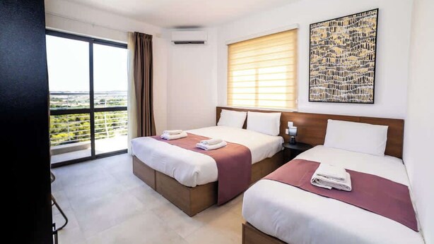 Ein Hotelzimmer mit zwei Betten, einem großen Fenster mit Jalousien, einem Deckenventilator und Blick auf die Stadt.