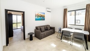 Ein modernes Hotelzimmer mit einer Couch, Esstisch, Stühlen, einem kleinen Beistelltisch und einem großen Fenster mit Vorhängen.