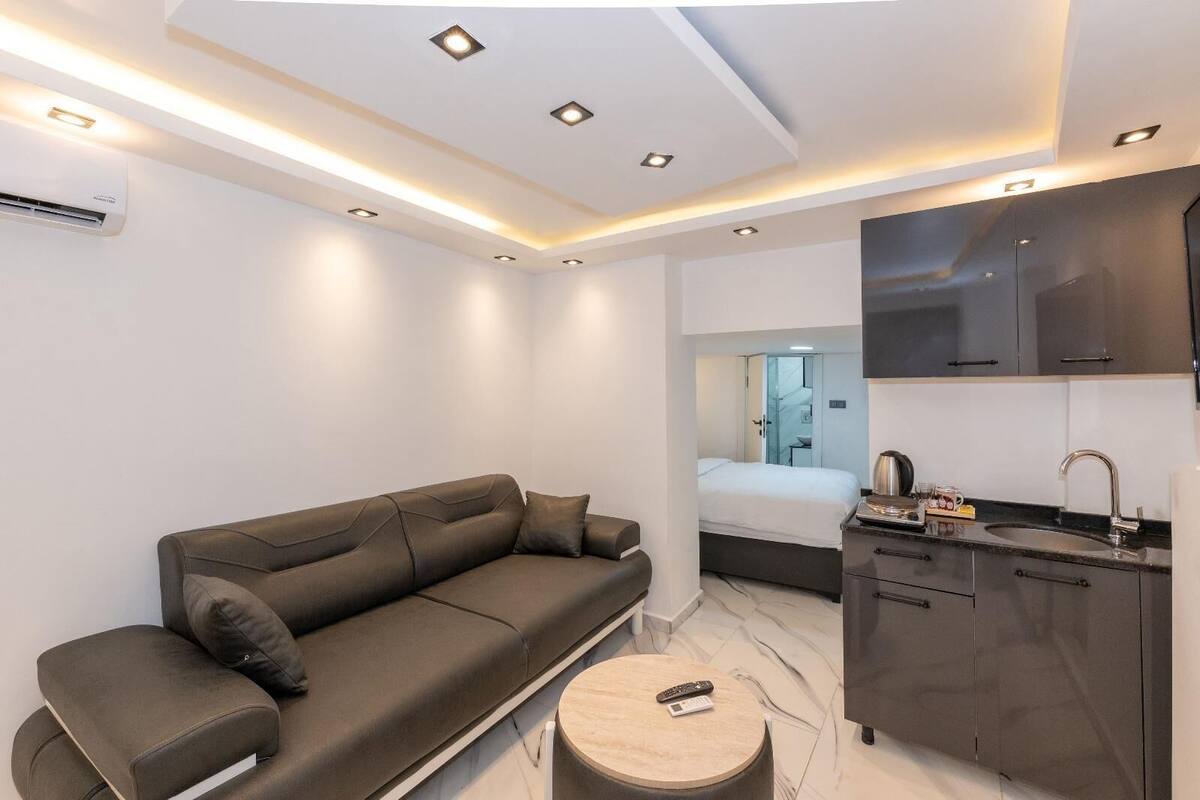 A modern hotel room with a sofa, a small round table, a bed, a kitchen area with a sink and cabinets, and a ceiling with recessed lighting.