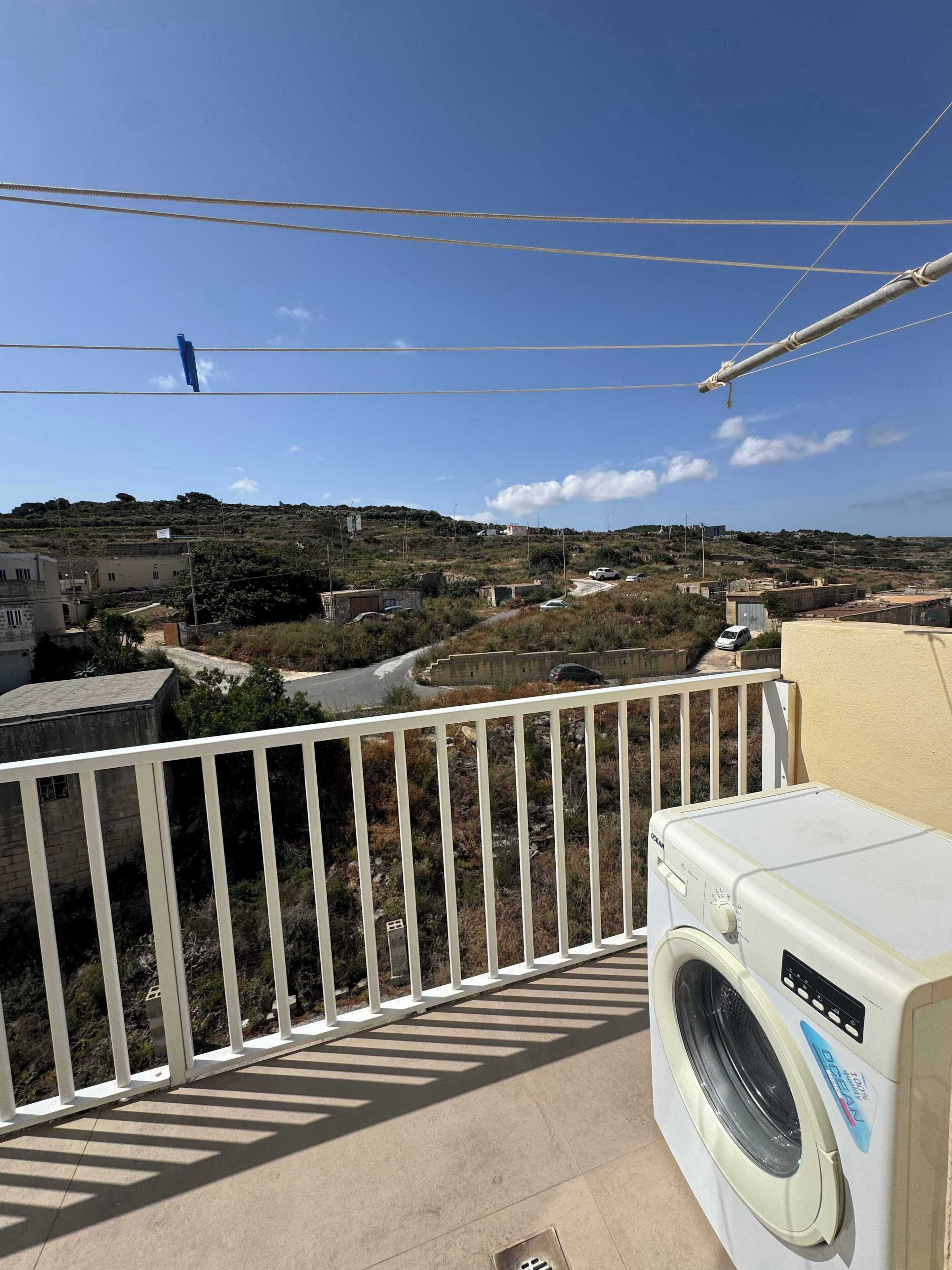 Een balkon met een wasmachine, een leuning en uitzicht op een glooiend landschap met gebouwen.