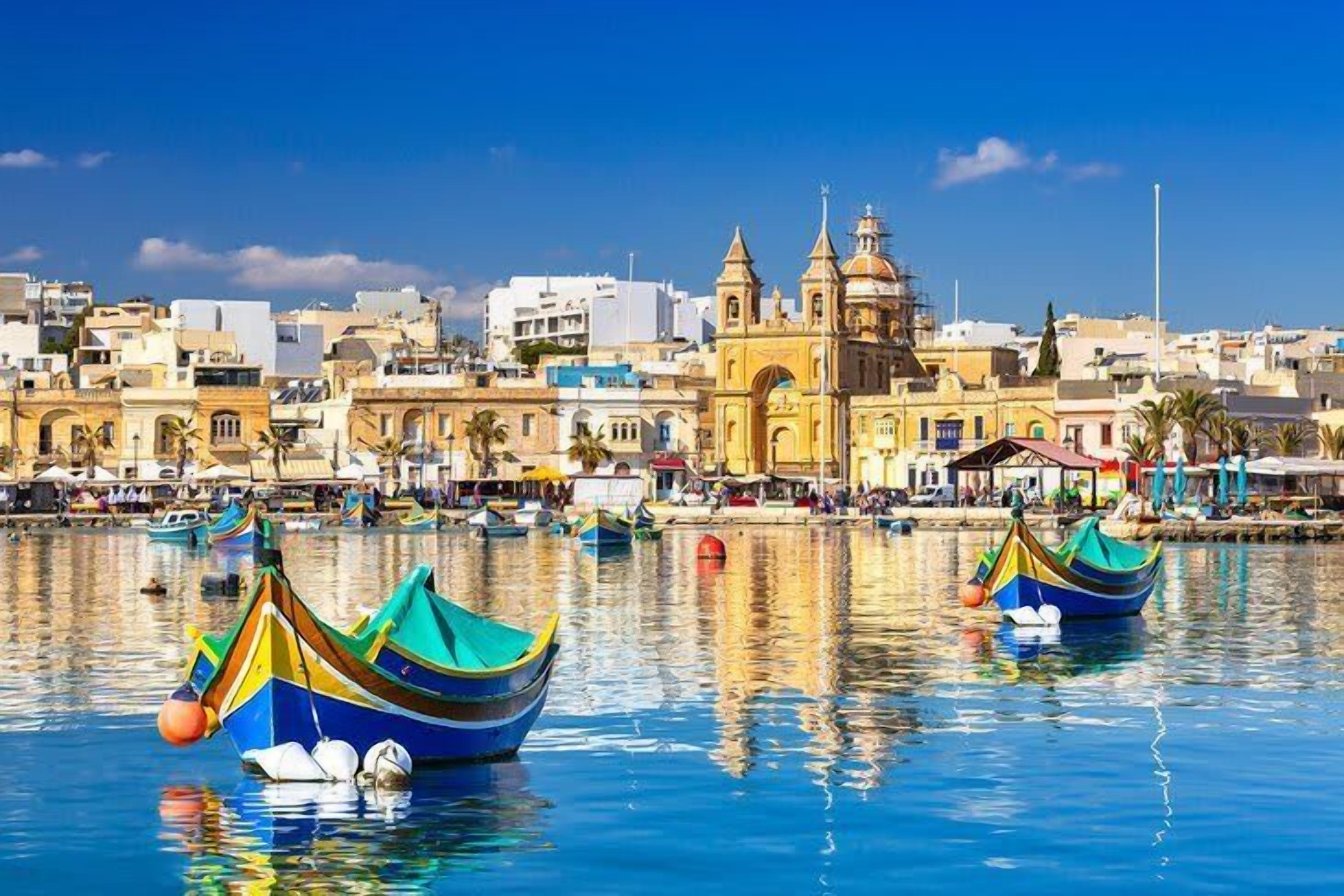 Una cittadina costiera con barche colorate ormeggiate nell'acqua, una chiesa importante con un campanile e un cielo azzurro limpido.