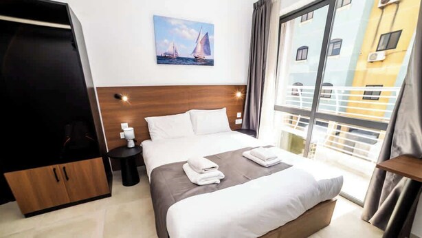 Quarto de hotel com uma cama, um guarda-roupa, uma mesa de cabeceira e vista para um edifício através de uma janela.