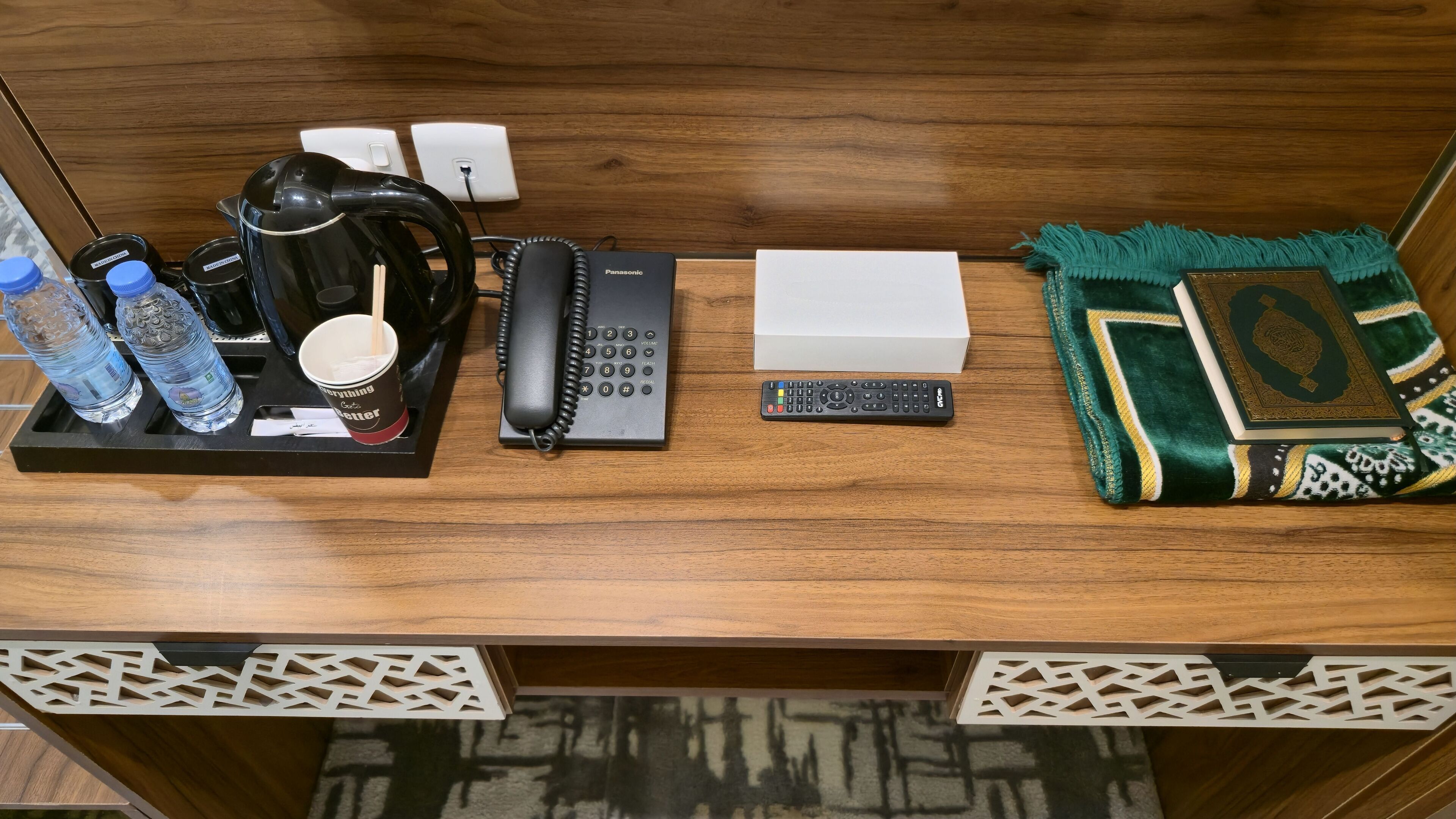 A wooden desk with a coffee maker, a telephone, a remote control, a water bottle, a cup, and a decorative book.
