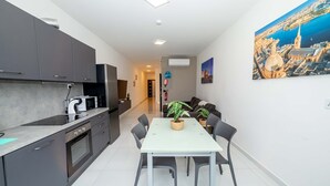 Een moderne keuken met grijze kastjes, een ingebouwde oven, en een eethoek met een witte tafel en stoelen. Er staan potplanten op de eettafel en er hangt een schilderij van een stadsgezicht aan de muur.