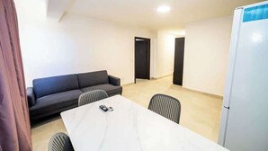 Una stanza con un tavolo bianco, due sedie, un divano scuro, un frigorifero bianco e una porta.