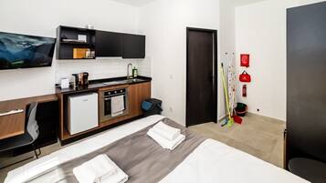 A modern hotel room with a bed, a small kitchenette, a flat-screen TV, and a door with a red sign.