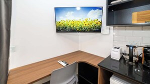 Una kitchenette moderna con un televisor de pantalla plana que muestra un campo de girasoles, una mesa de madera, una silla y una cafetera.