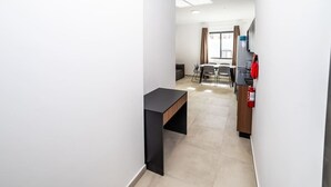 Un pasillo estrecho con un escritorio negro, un comedor con sillas y una mesa, y una zona de cocina con extintor.