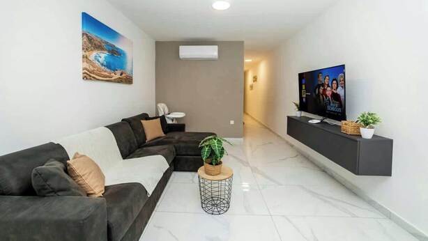 Una sala de estar moderna con un sofá modular, un televisor de pantalla plana, una planta en maceta y un cuadro de un paisaje costero.