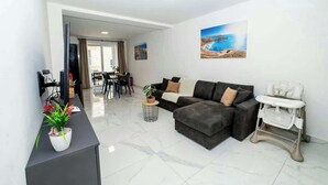 Una sala moderna con un sofá seccional gris, un comedor con mesa y sillas, y una pared blanca con un cuadro de una escena costera.
