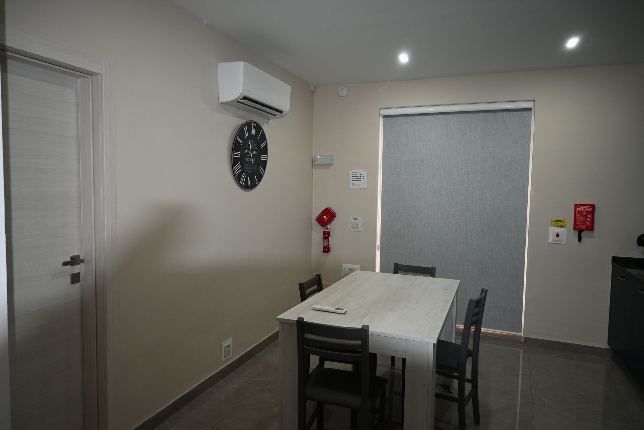 A room with a white table, four chairs, a clock, an air conditioner, a fire extinguisher, and a door.