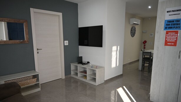 A modern living room with a flat-screen TV, a white door, a small white shelf, and a dining area with a chair and table. - Comfortable 3-Bedroom Seaside Stay (St. Paul's Bay)