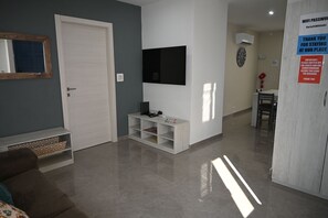 Una sala de estar moderna con un televisor de pantalla plana, una puerta blanca, un estante pequeño blanco y un comedor con una silla y una mesa.