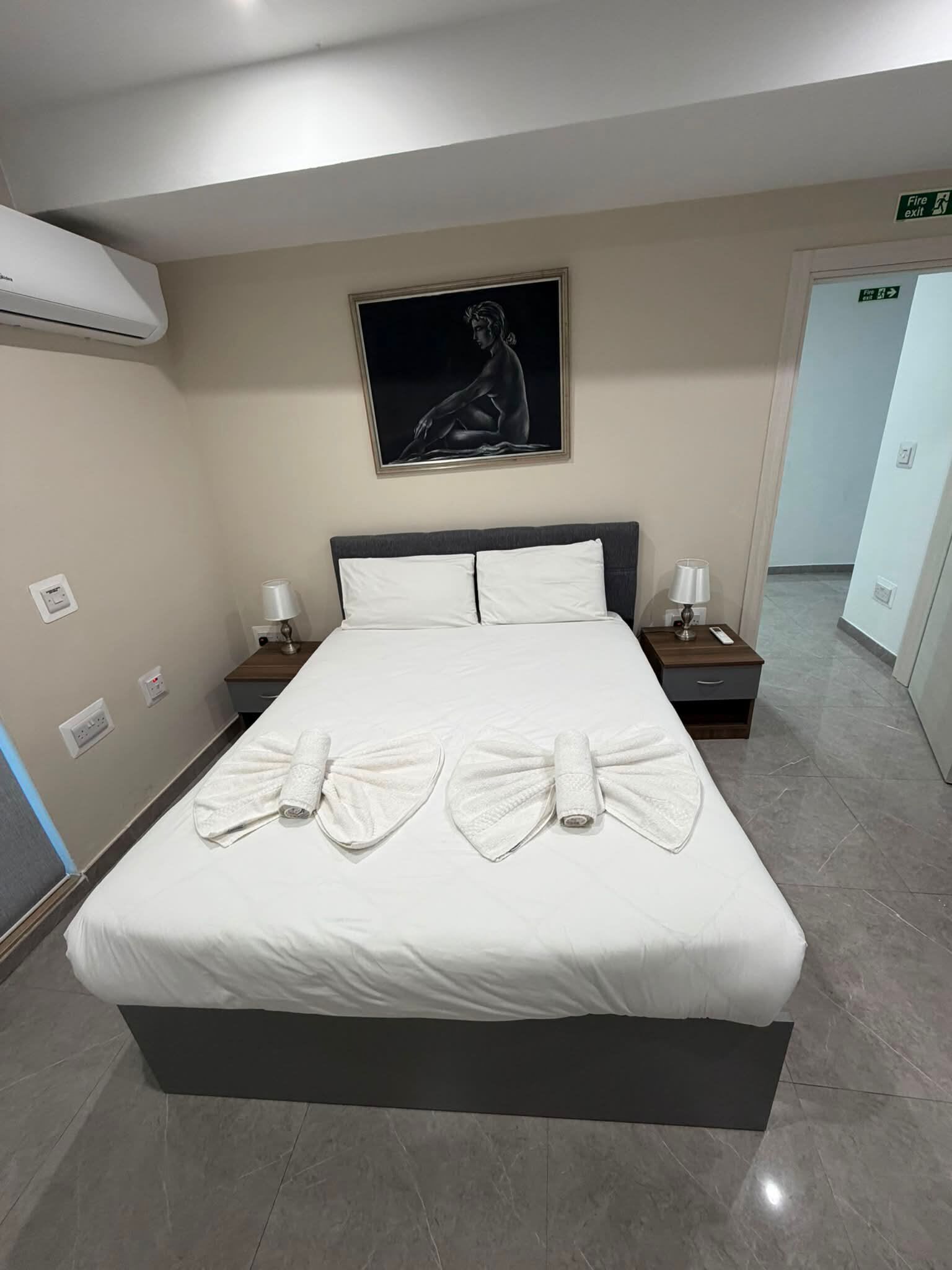 A hotel room with a large bed, two bedside tables with lamps, a painting on the wall, and a fire exit sign.