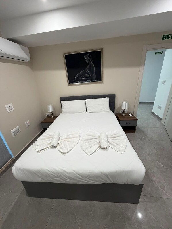 Una habitación de hotel con una cama grande, dos mesitas de noche con lámparas, un cuadro en la pared y un letrero de salida de incendios.