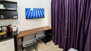 Une pièce compacte avec une kitchenette, un bureau en bois avec deux chaises, un téléviseur à écran plat fixé au mur et un rideau violet.