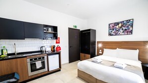 Une chambre d’hôtel moderne avec une kitchenette, un lit recouvert de draps blancs et un tableau accroché au mur.