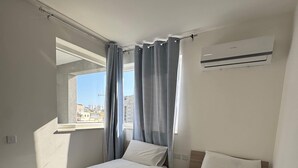Ein Hotelzimmer mit zwei Betten, einem Fenster mit Vorhängen, einer Klimaanlage und Blick auf Gebäude.