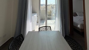 Ein Zimmer mit einem großen Tisch, zwei Stühlen, einem Balkon mit Blick auf Gebäude und einem Bett mit Holz Kopfteil.