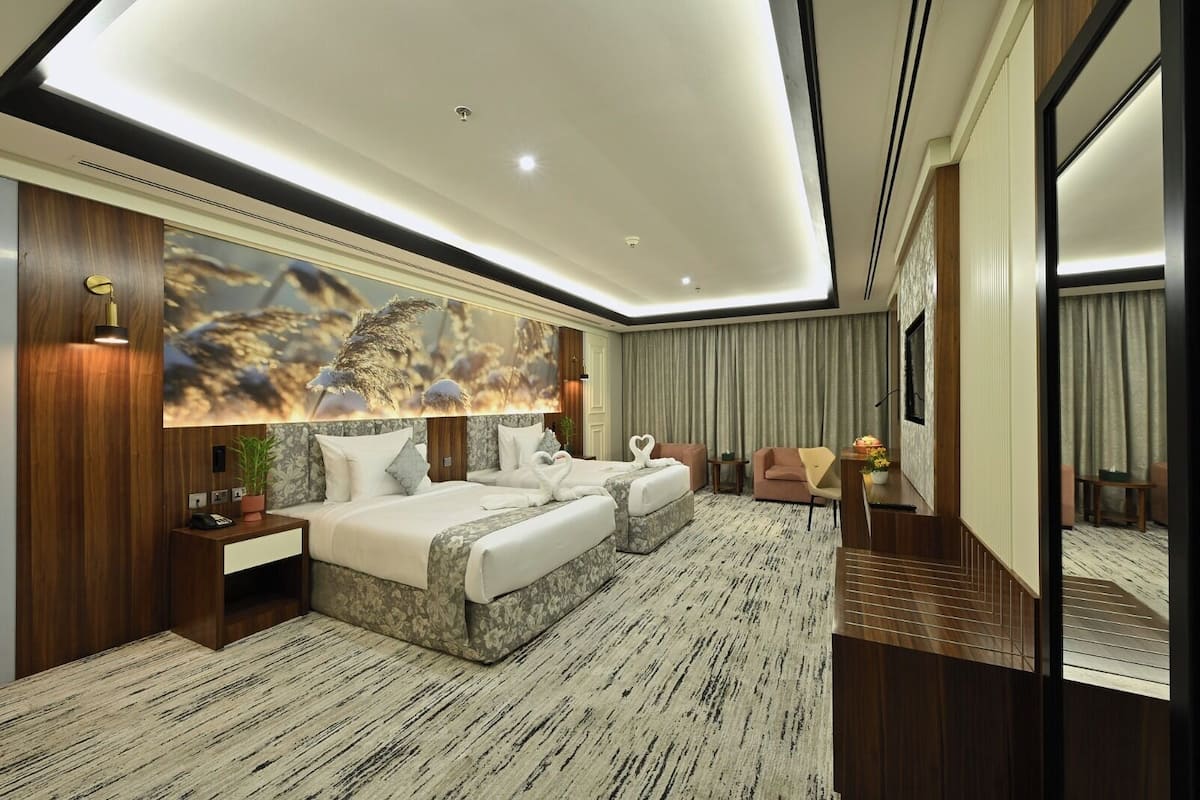 A modern hotel room with a large bed, a sitting area with a sofa and armchairs, a small table, and a large mirror.