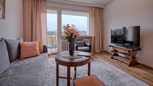 A modern living room with a sofa, a coffee table, a TV on a wooden stand, and a view of the outdoors through large windows.