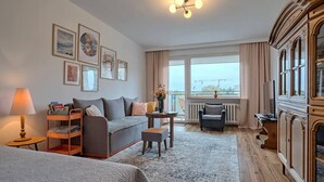 A hotel room with a sofa bed, a small table with a vase, a chair, a cabinet, and a view of the outside.