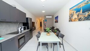 A modern kitchen with grey cabinets, a white countertop, and a dining area with a white table and chairs. There is a painting of a cityscape on the wall. - Spacious 2BR Apartment with Terrace – by HostMT (San Pawl il-Baħar)
