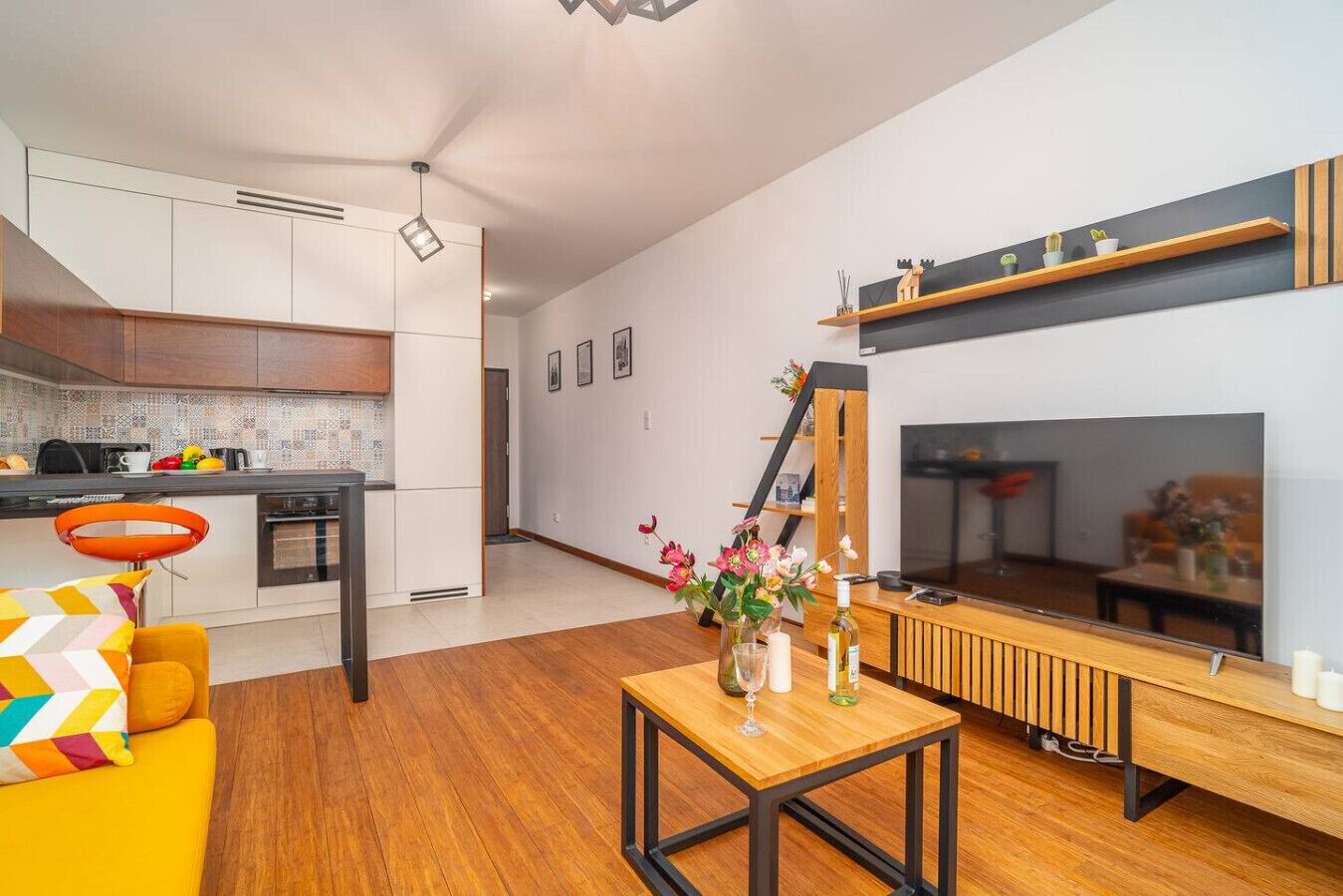 Un salon moderne avec un plancher en bois, un canapé jaune, une table basse agrémentée d’objets décoratifs, une cheminée et un coin cuisine doté de meubles blancs et d’un dosseret carrelé.