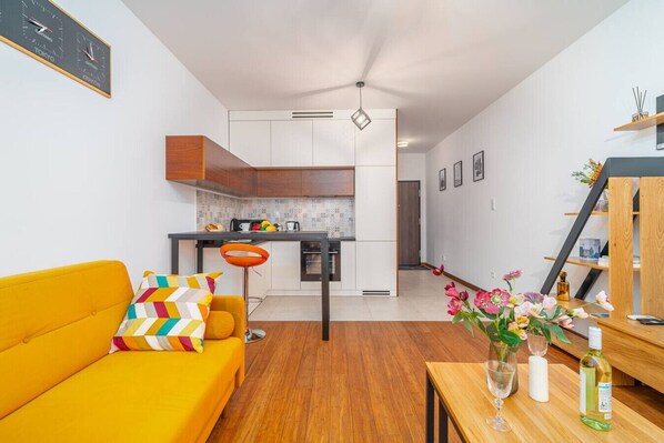 En moderne lejlighed med en lys gul sofa, et lille køkken med spiseplads, og et træspisebord med en vase blomster.