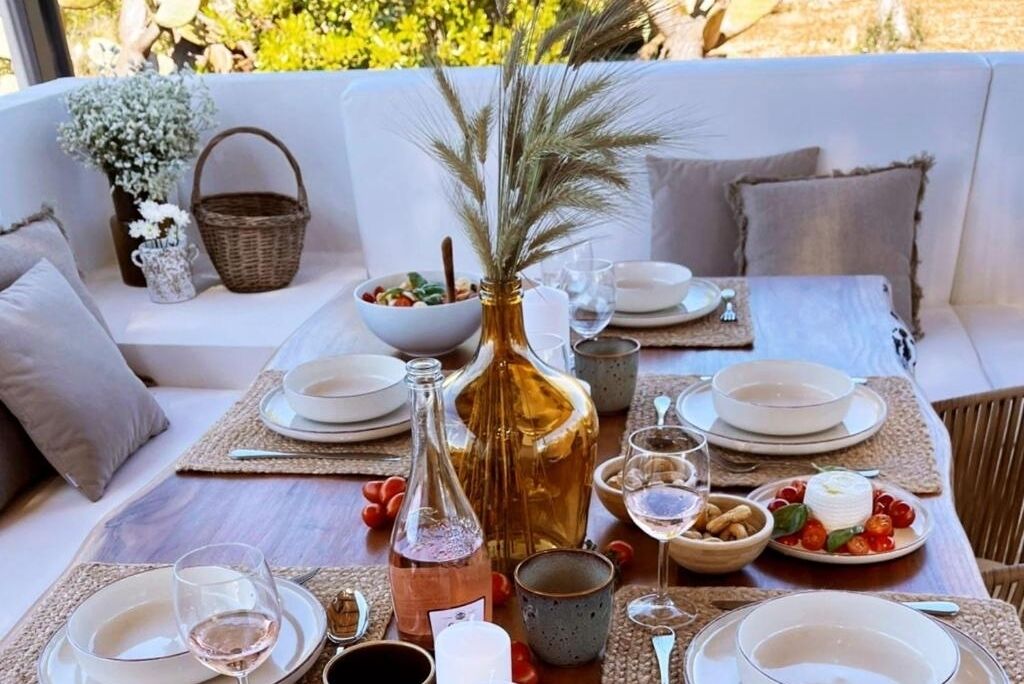 A dining table set with plates, glasses, and a bottle of wine, surrounded by cushioned seating and a vase with dried plants.