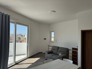 Una habitación moderna y minimalista con una puerta de cristal corredera que da a un balcón, un sofá gris, una mesita y vistas a la ciudad.