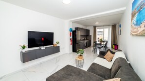 Uma sala de estar moderna com uma televisão de tela plana, sofás cinzas, uma mesa de centro e uma cozinha ao fundo.