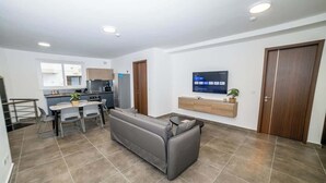 Un salon moderne avec un canapé gris, une table à manger avec des chaises, un petit coin cuisine et un téléviseur fixé au mur.