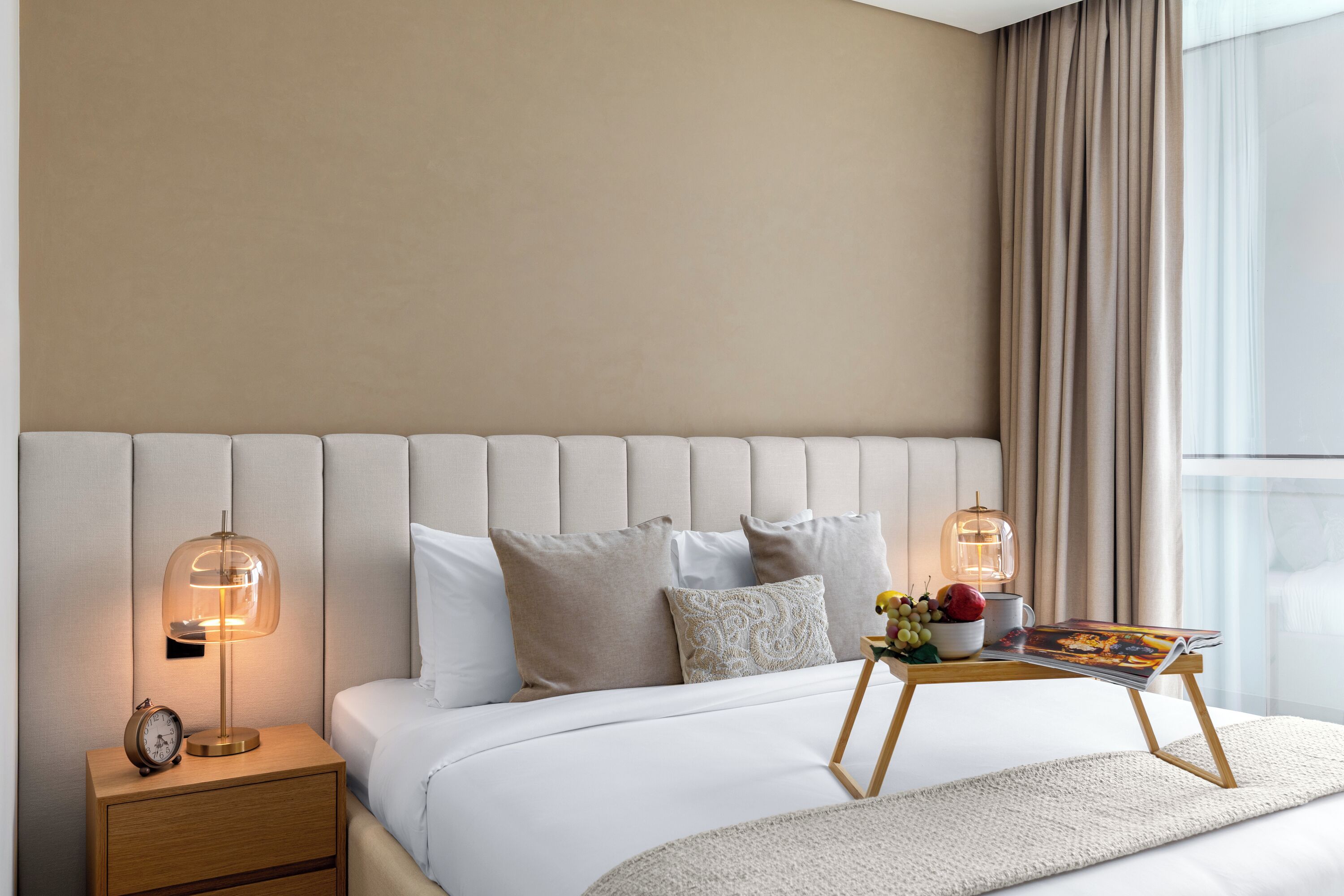 A neatly made bed with a headboard, bedside lamps, a bedside table with a clock, a magazine, and a fruit bowl.