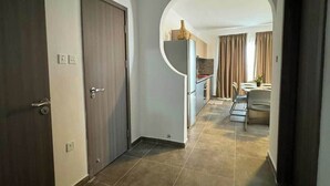 A hallway leading to a kitchen and dining area with a modern design, including a dark door, a light-colored wall, and a tiled floor. - Coastal Comfort Apartment – by HostMT (San Pawl il-Baħar)
