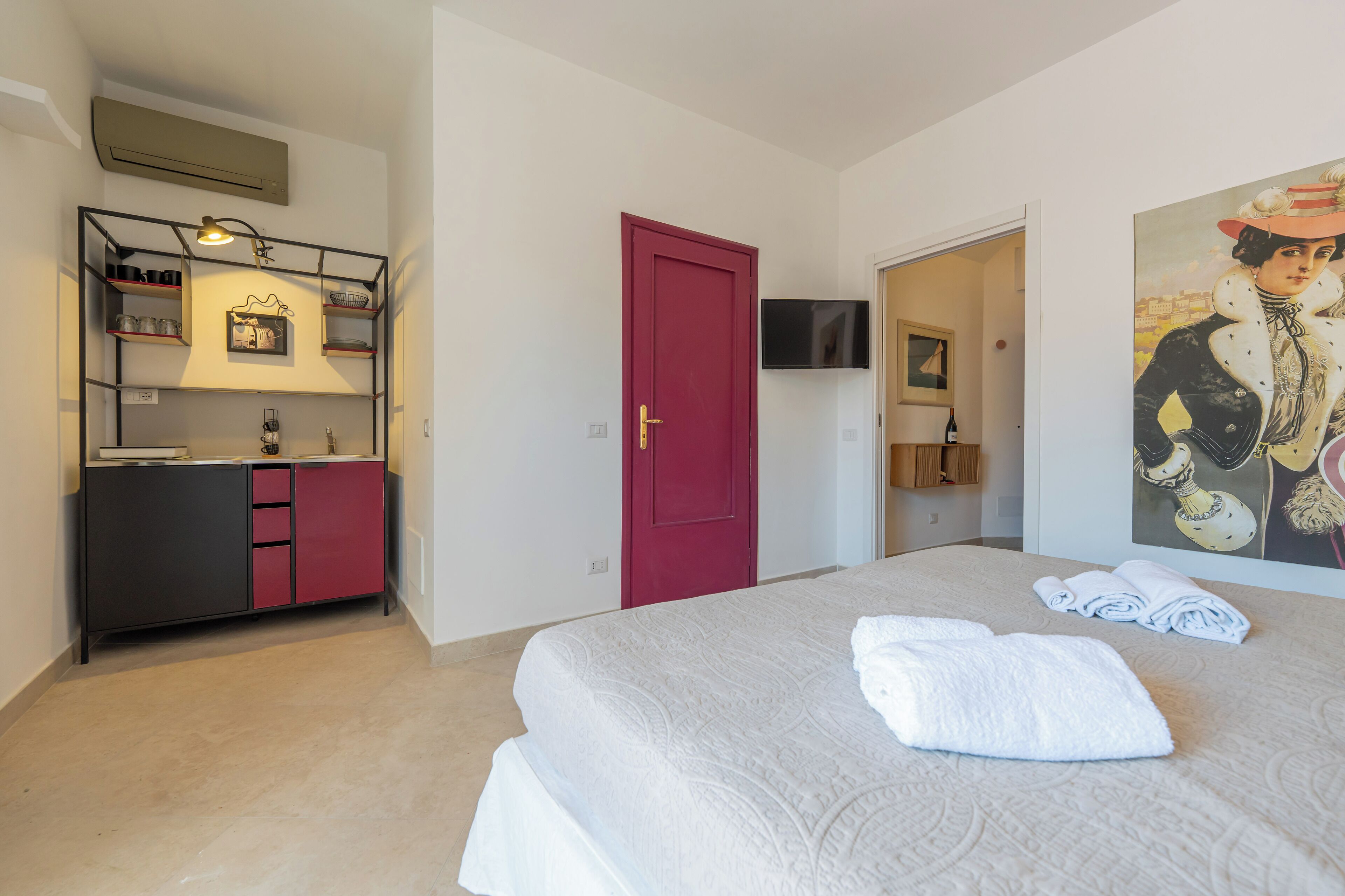 A hotel room with a bed, a red door, a TV mounted on the wall, and a painting.