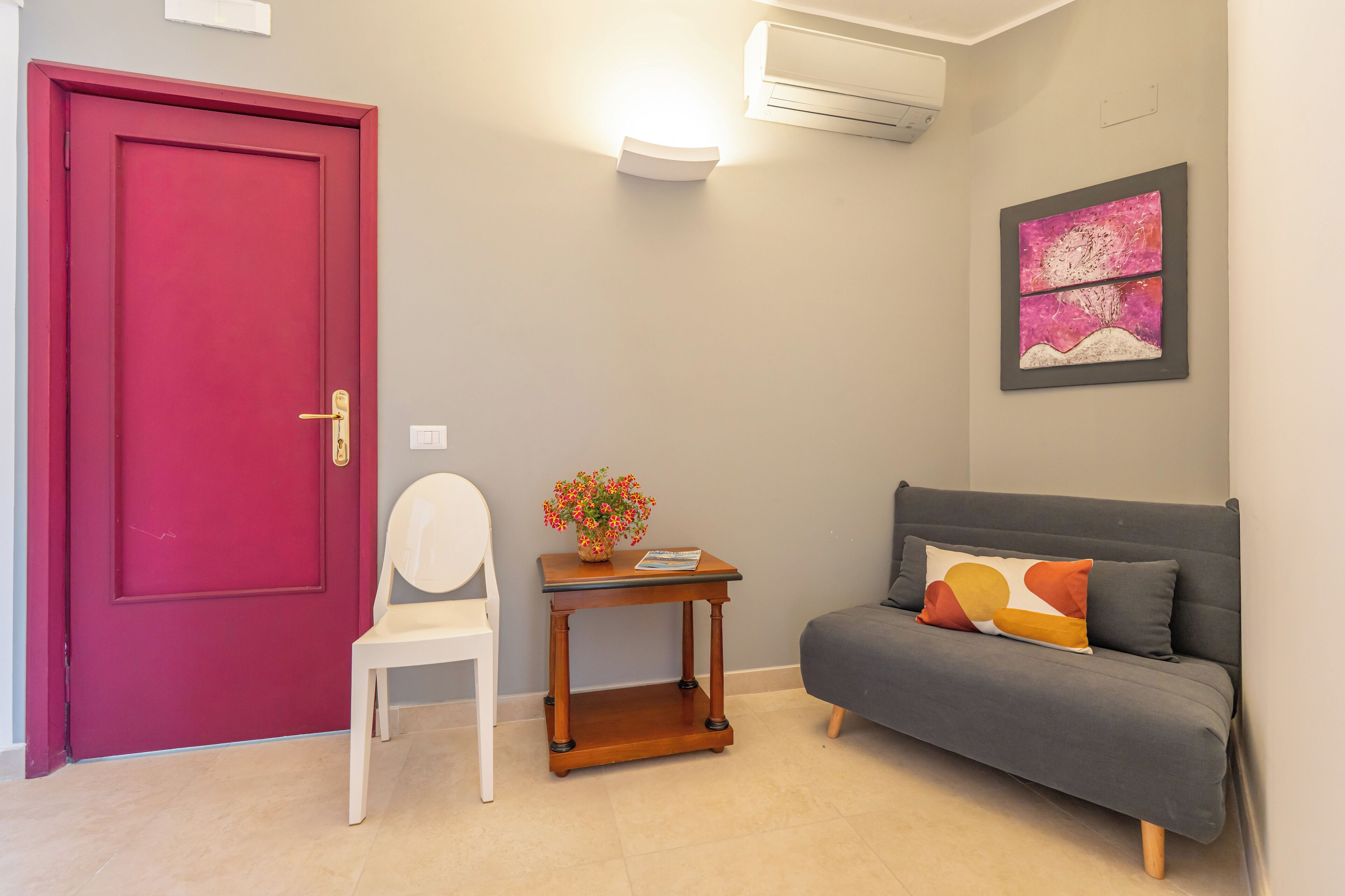 Une pièce avec une porte rouge, une chaise blanche, un canapé gris, une petite table en bois ornée d’un vase de fleurs et un climatiseur fixé au mur.