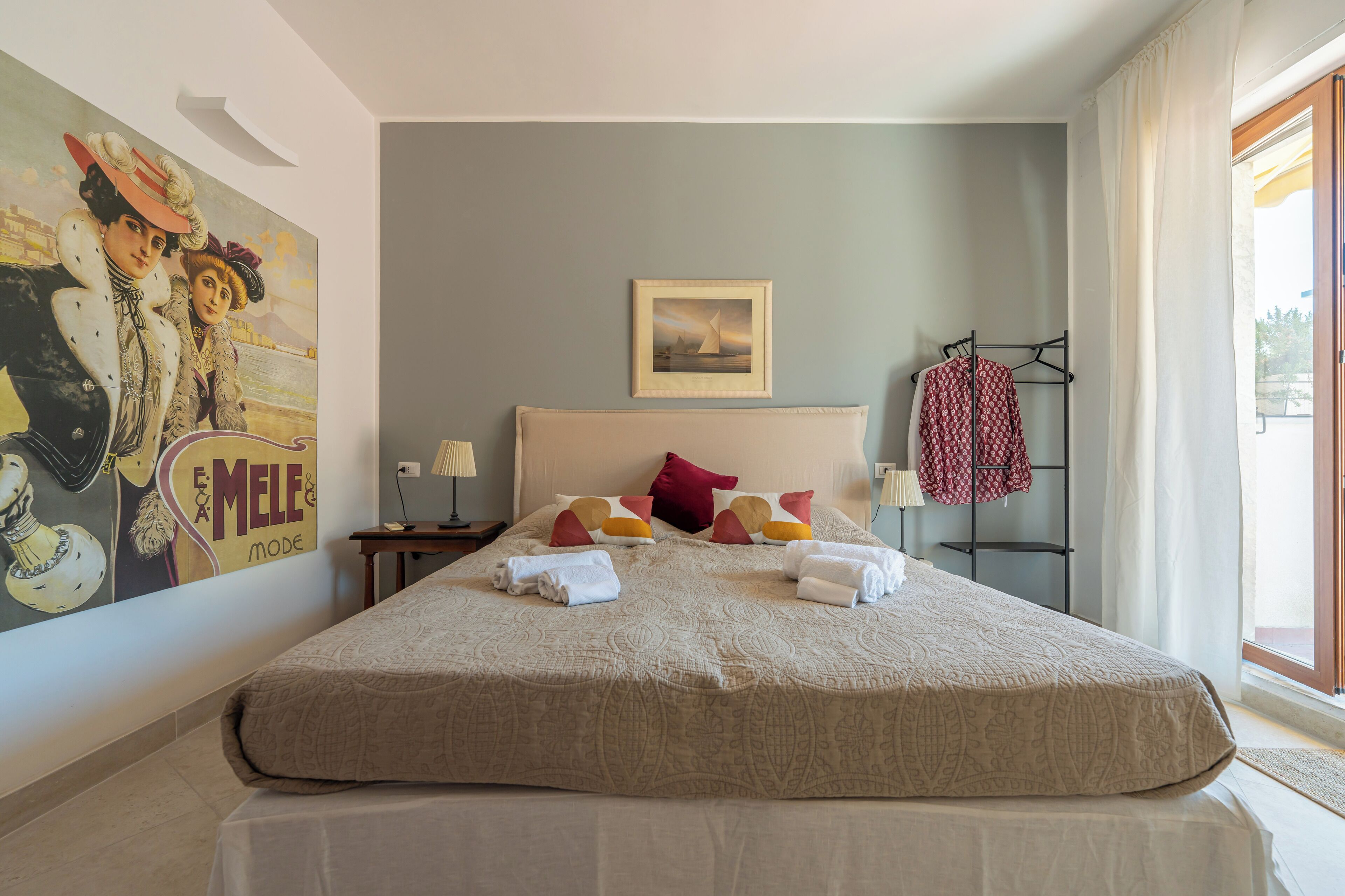 A neatly made bed with a beige bedspread and pillows, a framed picture above it, and a window with curtains on the right.
