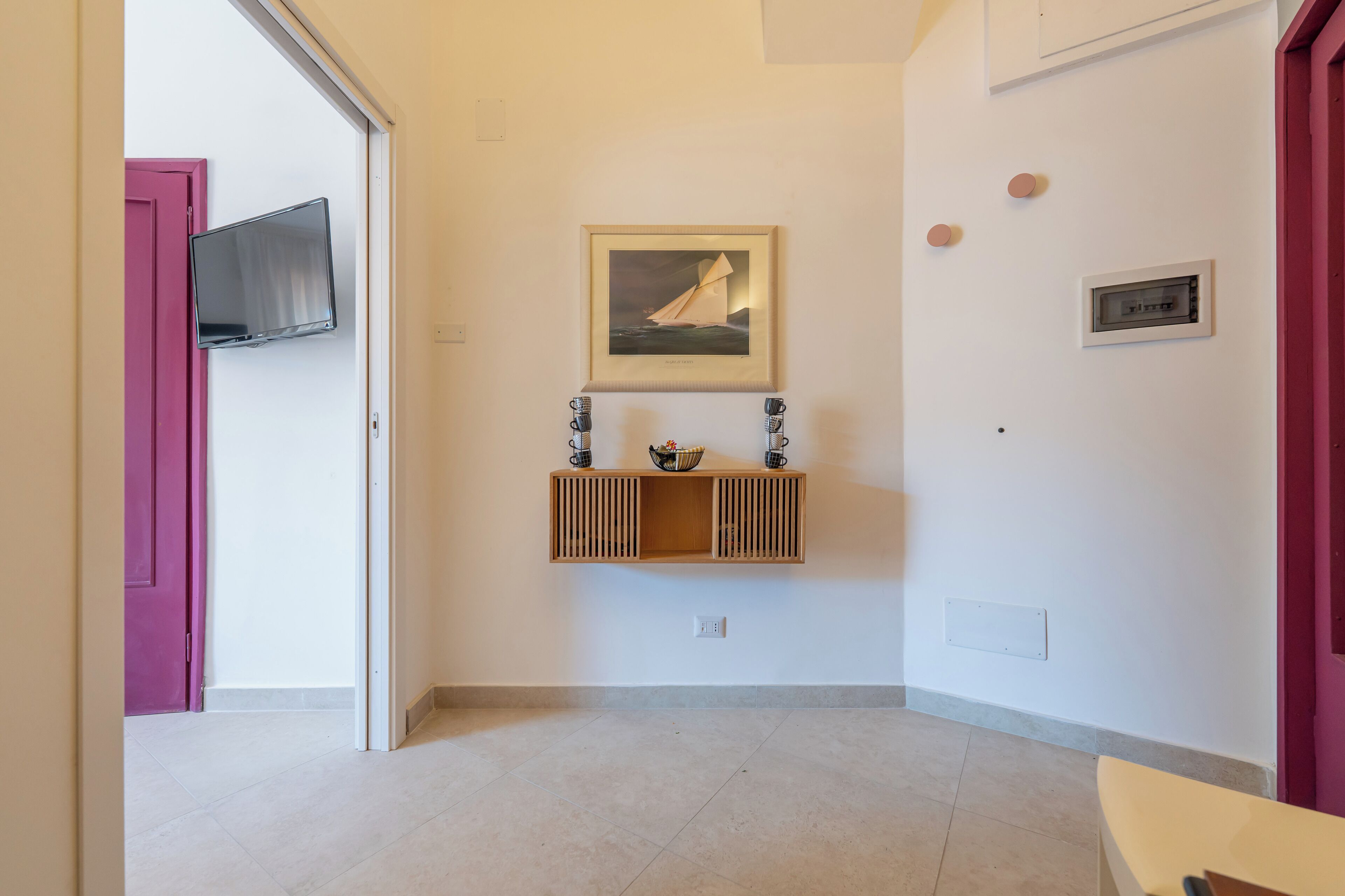 A hallway with a wooden console table, a framed picture, a mounted TV, and a door with a pink frame.