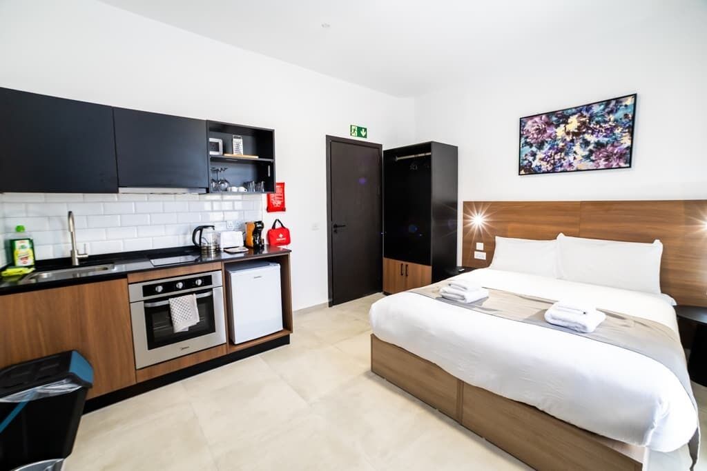 A modern hotel room with a kitchenette, a bed with white linens, and a painting on the wall.