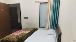 A single bed with white linens, a patterned blanket, and a wooden headboard. There is a window with curtains, a door, and a small air conditioning unit on the wall. - HOTEL K K PALACE (Khatoo)